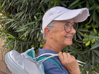 Active senior woman in white hat and eyeglasses walking outdoors holding her shoes on shoulders...