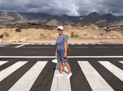 Active senior woman walking on the crosswalk holding her shoes in hand for her sore feet. Mountains and cloudy sky in the background - Powered by Adobe