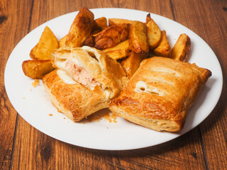 Two crispy cooked salmon pie and golden wedges on a white plate and wooden table. Tasty simple dinner serving. Fried fine products. Pastry filled with a fillet of a fish and creamy sauce