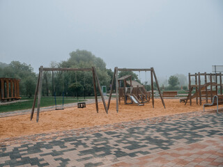 Modern playground on a sand surface and built with use wooden organic materials. No people, fog in the background. Concrete footpath around the play ground area.