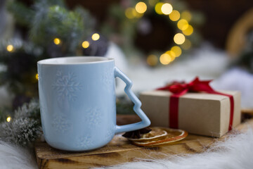 A blue mug of tea with marshmallows and lemon slices, cinnamon sticks, star anise. Christmas tree branches with lights