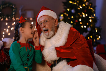 Shocked Santa Claus and cute little elf telling her wish on sofa in decorated room for Christmas at night