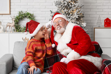 Santa Claus and cute little girl telling her wish on sofa in decorated room for Christmas