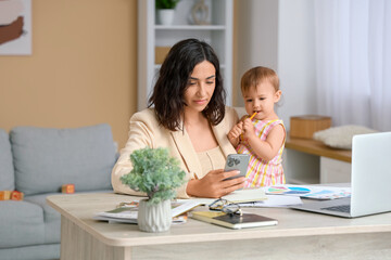 Cute little baby and young mother working with laptop while using mobile phone on maternity leave...