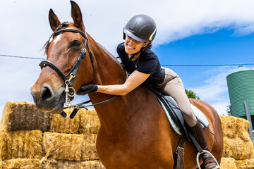 Smiling female rider hugging a brown horse near hay bales