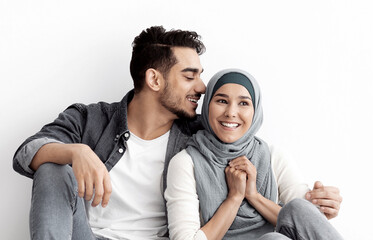 Joyful young woman in hijab sitting with her loving husband on floor in new empty apartment, dreaming about prosperous future of her family, white wall background, panorama with copy space