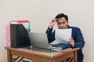 Shocked Southeast Asian Man Works while Sitting in front of a laptop and holding paperworks at Home office. The Workplace of a Professional Worker, Freelancer or Student. Distance Work Concept.