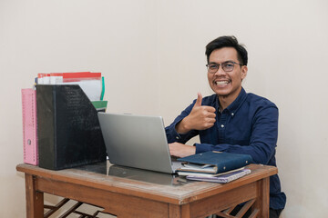 Smiling and happy Southeast Asian Man Raising thumbs up, Working While Sitting In Front Of Laptop In Home Office. Workplace Of Professional Worker, Freelancer or Student. Remote Work Concept