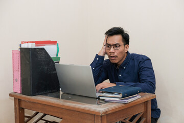 Shocked Southeast Asian Man Works while Sitting in front of a laptop at Home office. The Workplace of a Professional Worker, Freelancer or Student. Distance Work Concept.