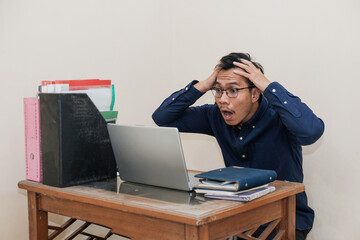 Shocked Southeast Asian Man Works while Sitting in front of a laptop at Home office. The Workplace of a Professional Worker, Freelancer or Student. Distance Work Concept.