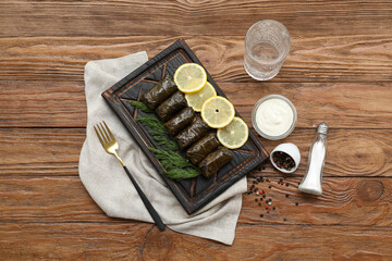 Cutting board with tasty Dolmas (stuffed grape leaves), sour cream, spices and glass of water on wooden background