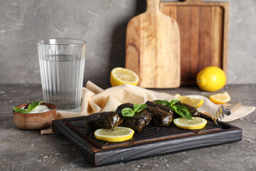 Wooden board with tasty Dolmas (stuffed grape leaves), lemons, sour cream and glass of water on grunge background