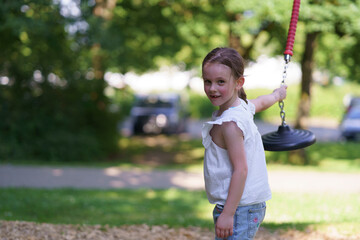 Little cute girl without front tooth rides on a suspended slide in a rope park in summer. Concept of an active healthy cheerful child