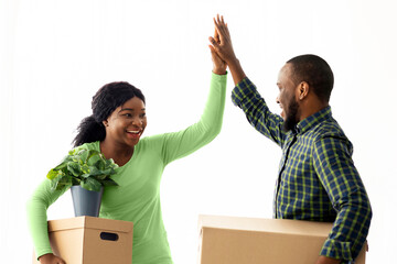 Happy Young Black Couple Giving High Five To Each Other, Celebrating Moving To New House. African American Spouses Carrying Cardboard Boxes And Clasping Hands, Enjoying Relocation, Free Space