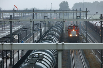 Rainy day at a train station.  Tank cars and a red locomotive are visible on the tracks.  Overhead wires and support structures are prominent.