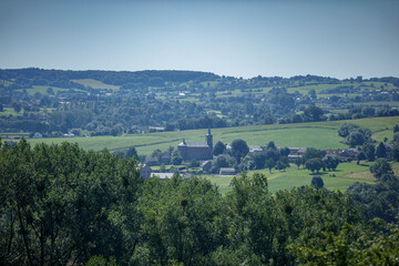 Naklejka premium Panoramic view of a rural European village nestled in a valley, with fields, trees, and a church steeple visible. Tranquil countryside scene.
