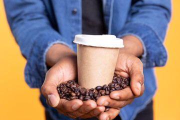 Male model holding a caffeine refreshment with black arabica coffee beans in his palms, standing against yellow background. African american man serving a cup of cold brew drink. Close up.