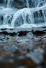 A cascading waterfall over rugged rocks, with every droplet of water and the moss-covered stones clearly visible. The surrounding greenery is lush, and the sunlight filters through the trees