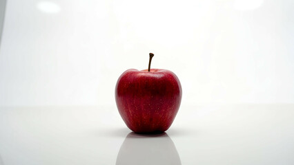 A single shiny red apple placed at the center of a stark white surface, showcasing its vibrant color and smooth texture under soft lighting.

