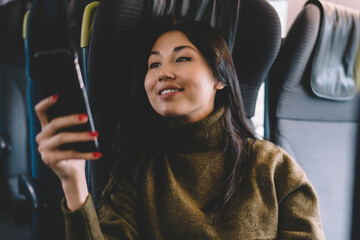 Positive asian female sitting in comfortable seat of locomotif traveling and using smartphone for chatting and checking social networks,smiling woman traveler enjoying wireless connection in train