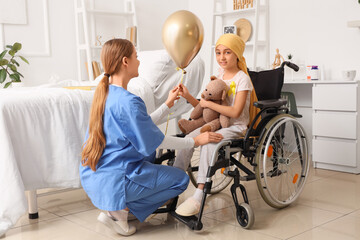 Little girl after chemotherapy in wheelchair taking golden balloon from nurse at hospital. Childhood cancer awareness concept