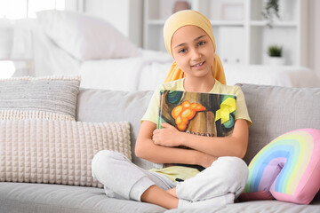 Little girl after chemotherapy with yellow ribbon and book sitting on sofa at hospital. Childhood cancer awareness concept