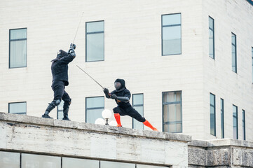 Two fencers dressed in black protective uniforms, helmets with face masks are fencing on castle...