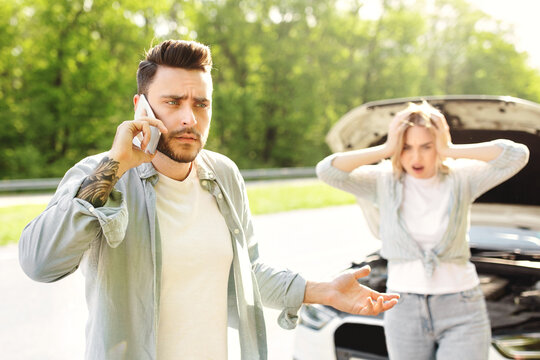 Young guy standing near his broken car together with terrified girlfriend, experiencing auto accident, calling breakdown road service, having difficulty receiving help in countryside