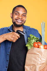 African american person using checkout scanner on bio produce in a paper bag, scanning natural homegrown veggies and promoting the local farming. Young male model with a device.