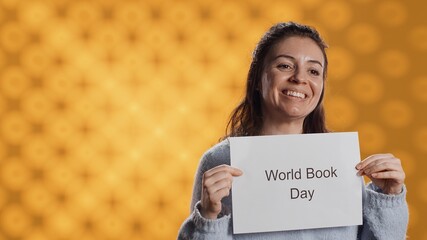 Smiling book club member holding placard with world book day message, isolated over studio background. Radiant bookworm recording video promoting literacy importance, camera A
