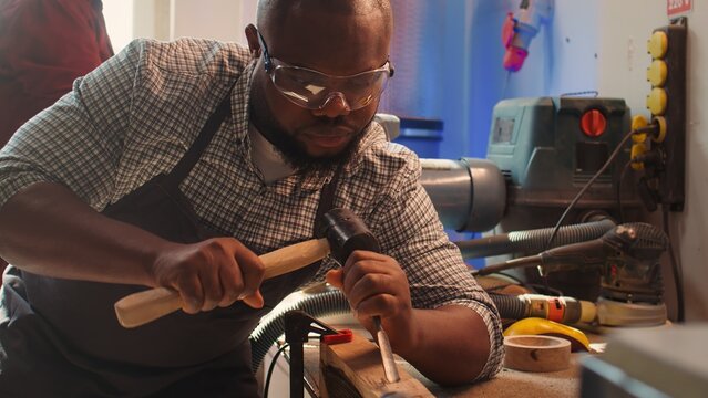 African american sculptor shaping raw timber using chisel and hammer in carpentry shop, creating wood art, wearing safety glasses. Artist making wood sculptures, engraving lumber with tools, camera B