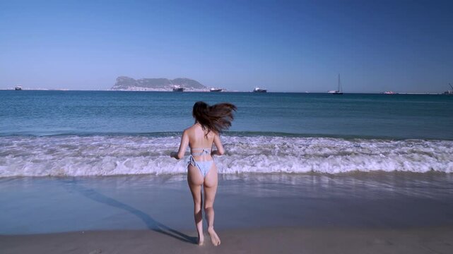 Chica joven corre sobre la arena de la playa hacia el mar, esta muy alegre por su sensacional d&iacute;a de sol y ba&ntilde;o en verano de vacaciones y lo expresa jugando con el agua, dando vueltas y una sonrisa.