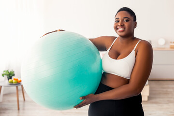 Fototapeta premium Portrait of plus size Afro lady in sportswear holding fitness ball after domestic training, indoors. Overweight black woman exercising at home, doing weight loss workout