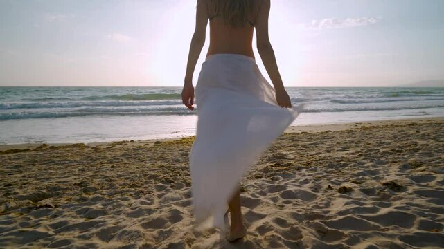Chica caminando por la playa hacia el mar al atardecer, con el sol y su estela brillante sobre el agua, con un pareo blanco transparente y expresando que esta muy a gusto con su sensacional d&iacute;a.