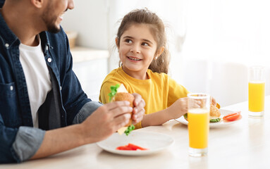 Portrait Of Happy Cute Little Arab Girl Eating Sandwiches With Father In Kitchen, Cheerful Female Child And Her Middle Eastern Father Sitting At Table, Enjoying Tasty Food And Drinking Orange Juice