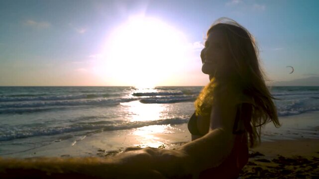 Chica Joven lleva a su amigo cogido de la mano hacia el mar al atardecer en la puesta de sol, mir&aacute;ndole y sonriendo muy contenta y alegre por el maravilloso d&iacute;a de sol y playa en verano de vacaciones.