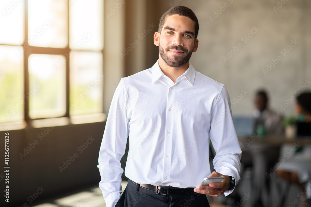 Poster a confident man in a white shirt holds a smartphone while interacting with his diverse team in a cor