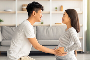 Obraz premium Young asian man helping his beautiful pregnant wife exercising, sitting together on fitness mat in living room. Expecting family practicing yoga at home. Sport and pregnancy cocnept