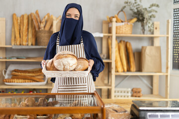 Muslim European girl works in sales area of bakery, selling bread in paper bag. She holds basket filled with fresh fragrant loaves of bread. Store employee near counter sells bakery products