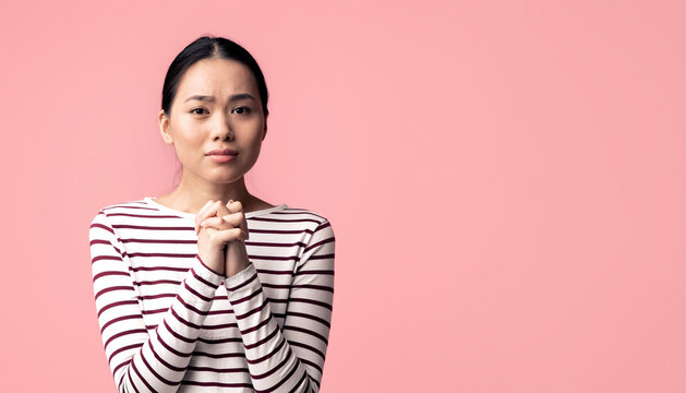 Please. Young asian female begging for something with clasped hands and looking at camera, desperate sad korean woman pleading and asking for help while standing over pink background, copy space
