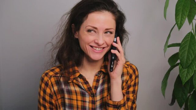 Happy laughing and smiling woman talking on mobile phone at home near the green frower in bright orange style shirt and texting on the screen to buy online. Closeup natural positive expression video	
