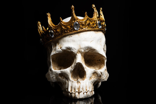An isolated front view of the skull of a king or queen monarch wearing a golden crown on a pain black background.