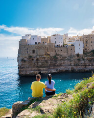 Polignano a Mare Italy, A couple sits together on the rocky cliff, gazing at the picturesque white...