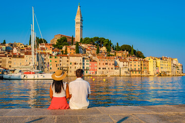 As the sun begins to set, a diverse couple of man and woman sits by the calm waters in Rovinj, Croatia, admiring the vibrant buildings and reflecting on the day's adventures. © Fokke Baarssen