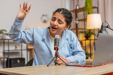 Young positive Indian woman blogger singer using laptop computer while singing into microphone and...