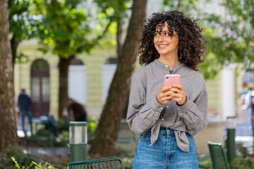 Fototapeta premium Caucasian young woman using smartphone typing text messages in social media application online, surfing internet, relaxing, taking a break outdoors. Lady girl standing on urban city street. Sunny park