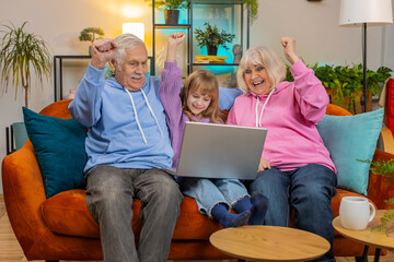 Surprise Caucasian grandfather, grandmother and granddaughter using laptop netbook while celebrating success at home. Happy girl with grandparents amazed by online game win clenching fists on sofa.