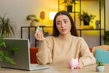 Poor Caucasian young woman insufficient amount of money, holding piggybank and one dollar banknote. Financial crisis. Bankruptcy. Poverty and destitution. Female girl sitting at home office at table