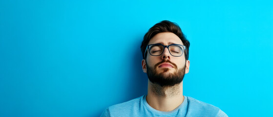 Obraz premium A young man with glasses relaxes against a vibrant blue wall, exuding calmness and tranquility.