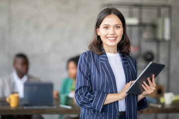 A confident woman stands in front of her diverse team in a contemporary office. She holds a tablet and smiles, as colleagues engage in a productive discussion while working together on projects.